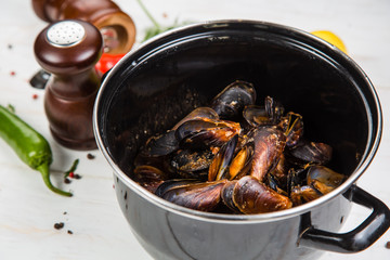 Spicy mussels in black saucepan. White wooden background decorated with lemon, hot peppers and greens