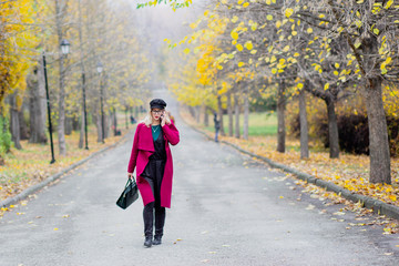 Cute young woman in glasses and cap, in a red coat walking in the park in the fall of the year