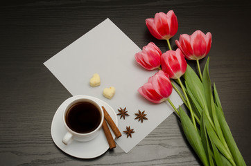 Holiday concept. Bouquet of pink tulips, a cup of coffee, heart-shaped sugar, cinnamon, star anise and sheet of paper on a black wooden background