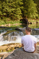 Man in white t-shirt sitting on the bank of the river against the background of a waterfall and green forest. Sunny day. Back view.