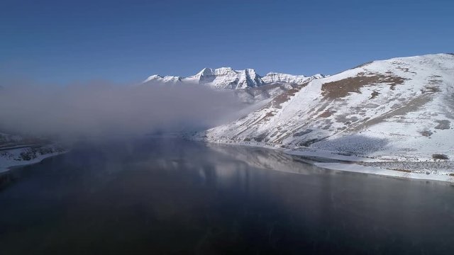 Aerial view flying over lake towards clouds in winter landscape rising up viewing snow covered Mount Timpanogos.