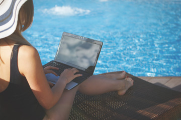Young cute woman in a beach hat is working with a laptop near the pool with blue water in the resort hotel. Back view.