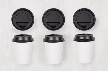 Coffee mockup - set of three white paper cups and blank black caps on white wood board, top view....