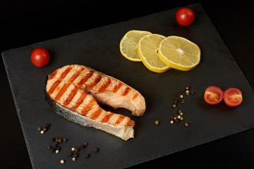 Grilled salmon steak with a pepper, lemon and tomatoes on a slate plate.