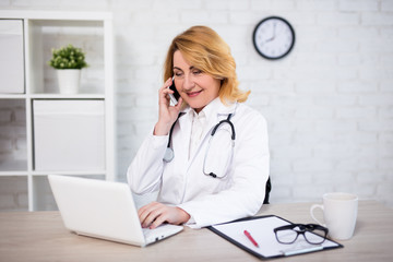 cheerful mature woman doctor or nurse working with laptop and talking by phone in office