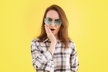 Fototapeta premium Photo of frightened Caucasian woman looks with bugged eyes, keeps mouth widely opened, witnesses terrible accident, has displeased facial expression, in a stylish shirt and glasses, yellow background