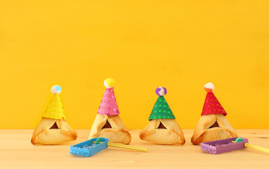 Purim celebration concept (jewish carnival holiday). Traditional hamantaschen cookies with cute clown hats and noisemaker over wooden table.