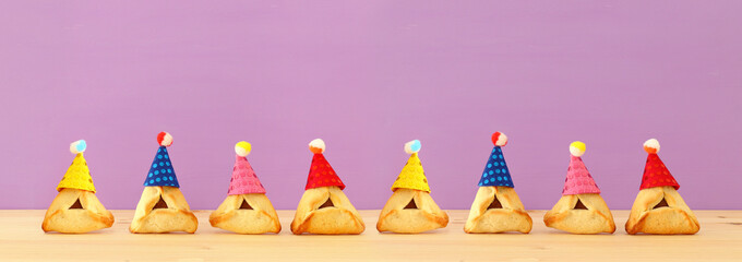 Purim celebration concept (jewish carnival holiday). Traditional hamantaschen cookies with cute clown hats over wooden table and purple background. Banner.