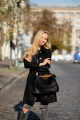 Street portrait of elegant smiling blonde model wearing black coat torn jeans walking at the city