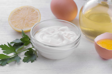 Obraz na płótnie Canvas Mayonnaise in a glass bowl and ingredients for making mayonnaise on a white wooden background. white sauce.