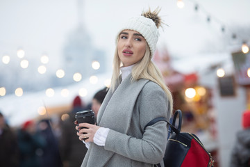 Fototapeta premium Happy winter time in big city of charming girl walking on street in coat with backpack. Enjoying snowfall, expressing positivity, smiling to camera, joyful cheerful mood, true emotions, new year mood.