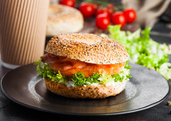 Fresh healthy bagel sandwich with salmon, ricotta and lettuce on black plate on black kitchen table background. Healthy diet food. Paper cup of coffee and fresh vegetables