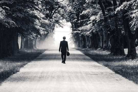 Black and white photo of a businessman or gentleman in a suit walking away on a dusty gravel road. Business man walking in an alley. Old trees and mysterious leaving of a man in a suit.