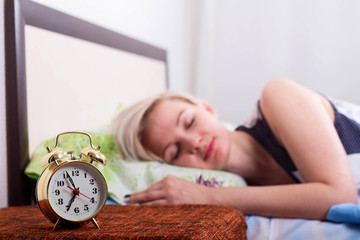 Close-up of alarm clock and sleeping young blonde woman in bright bedroom at home, morning.