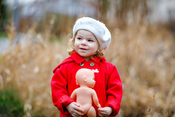 Outdoor portrait of little cute toddler girl in red coat and white fashion hat barret with toy...