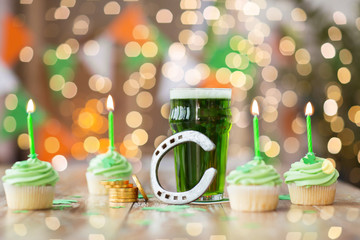 st patricks day, holidays and celebration concept - glass of green beer, cupcakes with candles, horseshoe and gold coins on wooden table