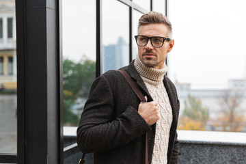 Portrait of stylish man 30s wearing eyeglasses, walking through city street near the building
