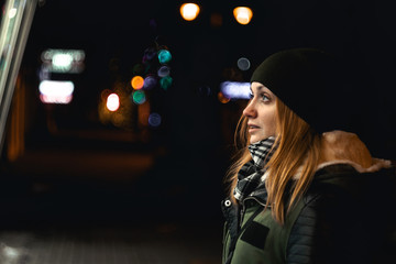 Outdoor night photo of young happy smiling girl looking through shop window, city lights on background, in street of european city, wearing knitted olive hat, scarf