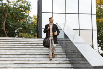 Image of bearded man 30s wearing eyeglasses, walking through city street and holding takeaway coffee