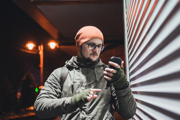 Portrait handsome young man with a beard and mustache use smartphone. glasses, and a hat. cold season, military style clothing. on the street in the city at night, scarf tube and olive gloves