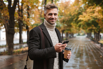 Photo of handsome man 30s wearing warm clothes walking outdoor through autumn park, and using mobile phone