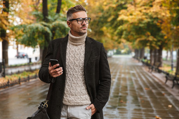 Photo of caucasian man 30s wearing warm clothes walking outdoor through autumn park, and using cell...