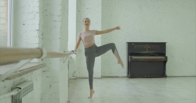 Elegant classic ballet dancer in sporty clothes rehearsing in ballet class. Beautiful ballerina with perfect body performing choreographic exercises, doing dehors at ballet barre in dance studio.