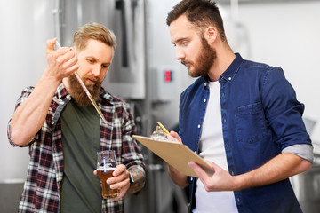 alcohol production, manufacture, business and people concept - male brewers with pipette and clipboard testing craft beer at brewery