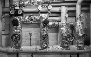 Black-and-white picture of heating pipes in the cellar with shut-off valves, thermometers, valves and throttles, jacket made of insulating wool and aluminium foil