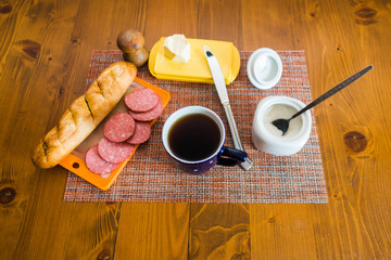 Breakfast: sliced sausage, baguette, butter, cup of tea and some sugar on the brown wooden table.