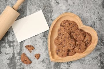 Chocolate chip cookies on heart-shaped plate with blank message card on grey stone background