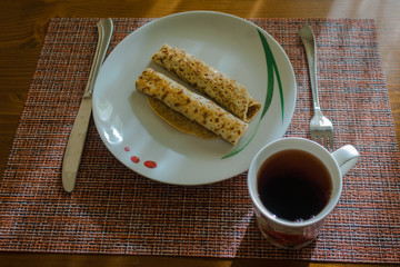 Two pancakes with meat on a large white plate and a cup of tea, served for breakfast on a dark wooden table. Shot in the morning under natural light from the window.