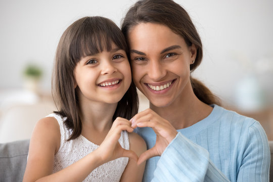 Cute little daughter and happy mother join hands in shape of heart as concept of mom and child love care support, smiling mum and her kid girl looking at camera posing together for headshot portrait