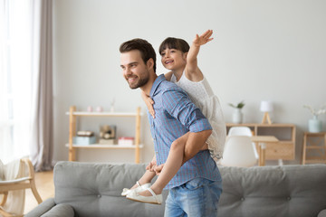 Happy dad piggybacking little funny daughter playing with daddy at home, cheerful single parent carrying kid on back having fun together, child girl enjoying active game with father in living room