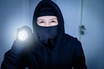 Porträt eines Einbrechers mit Sturmmaske und leuchtender Taschenlampe im dunklen Haus