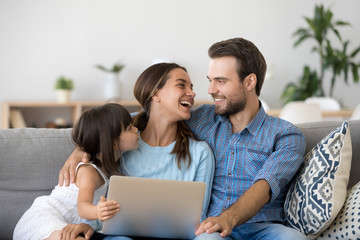 Happy family of three bonding using laptop sitting on couch at home, smiling parents with kid daughter having fun talking laughing relaxing with computer doing internet shopping together on weekend