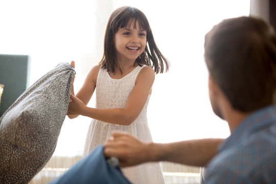 Cute little child girl laughing holding cushion having funny pillow fight with dad, happy joyful kid daughter playing active game with father enjoying spending fun time on weekend with daddy at home