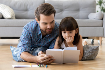 Cute little girl listening to dad reading fairy tale lying on warm floor together, caring father holding book teaching child daughter spending time with focused kid, family hobbies activities at home