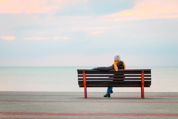 Mature woman, with beautiful long hair, wearing white knitted hat, dark coat and black gloves, sits on the bench admiring seascape. Winter mood. Copy space.