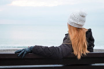 Mature woman, with beautiful long hair, wearing white knitted hat, dark coat and black gloves, sits on the bench admiring seascape. Winter mood. Copy space.