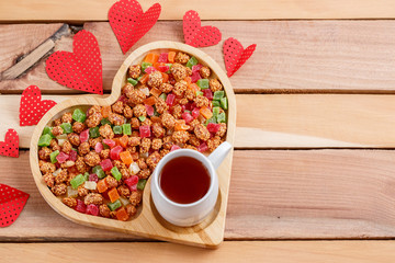 Dried fruits and nuts with tea, on a bamboo stand in the shape of a heart on a wooden table. Also nearby are various cardboard hearts and a box with a heart-shaped gift.