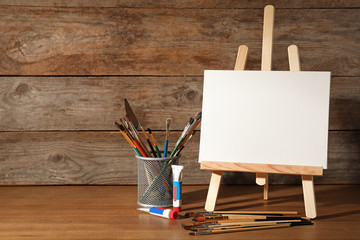 Easel with blank canvas board and painting tools for children on table near wooden wall. Space for...