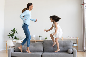 Happy mum and little daughter playing jumping on comfortable sofa in living room, mother baby...