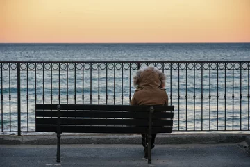 Fototapete Rund Woman with hooded parka sitting on a bench in front of the sea at sunset in winter, Alassio, Liguria, Italy © Simona Sirio