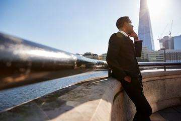 Young black businessman wearing a suit leaning on a wall at Thames embankment, London, using...