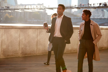 Two business colleagues walking by the Thames riverside in the city of London talking, man holding...