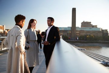 Side view of three millennial business colleagues stand talking on Millennium Bridge, London,...
