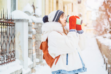 Gorgeous female drinking hot dring from the thermo cup on street in good mood. Outdoor photo of glad woman in knitted red mittens, white fur jacket having fun during winter photoshoot before christmas