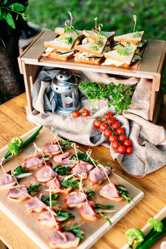 Delicious catering banquet buffet table decorated in rustic style in the garden. Different snacks, sandwiches with ham, chiken, cheese and greenery on a wooden plate. Outdoor. Vertical photo.