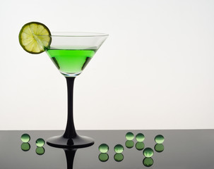 Martini glass with a green drink and a slice of lime, standing on a dark reflective table, next to the green transparent balls